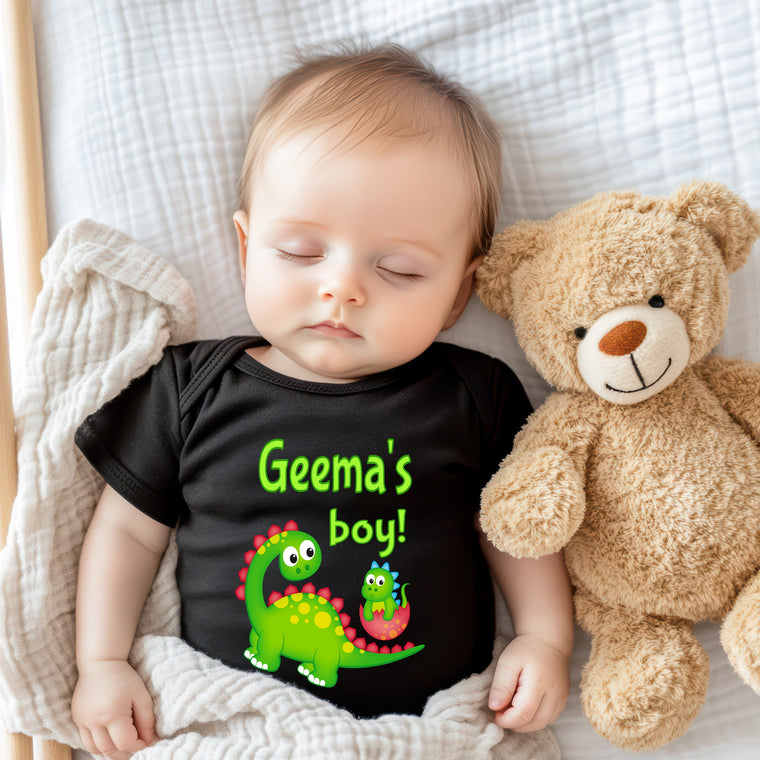 A sleeping baby wearing a black t-shirt with a dinosaur design and the text "Geme's boy!" next to a teddy bear.