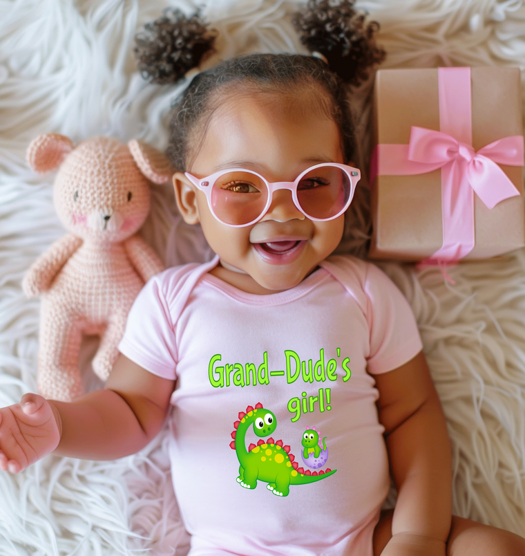 A smiling baby girl wearing a pink outfit with a dinosaur graphic and glasses, lying on a white surface next to a pink gift box.