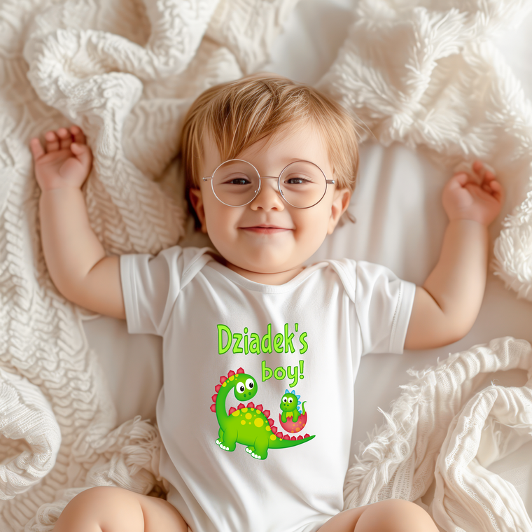 A smiling baby wearing a white onesie with a dinosaur graphic and the text "Dzia Dusk's boy!" lying on a white blanket.