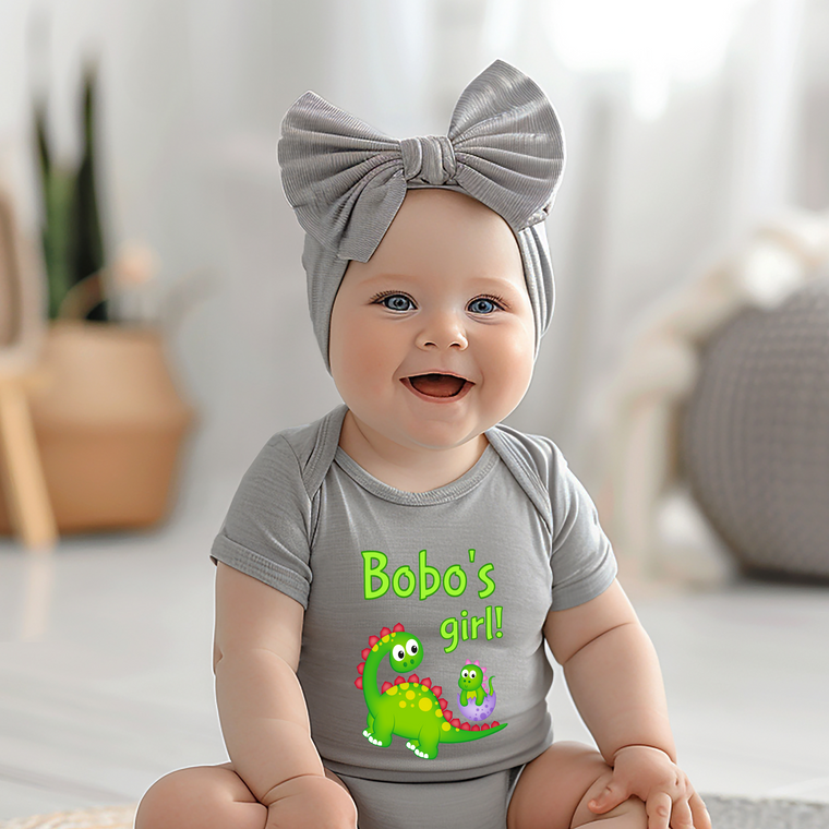 A smiling baby girl wearing a gray headband and a gray onesie with a dinosaur graphic, sitting on a white surface.
