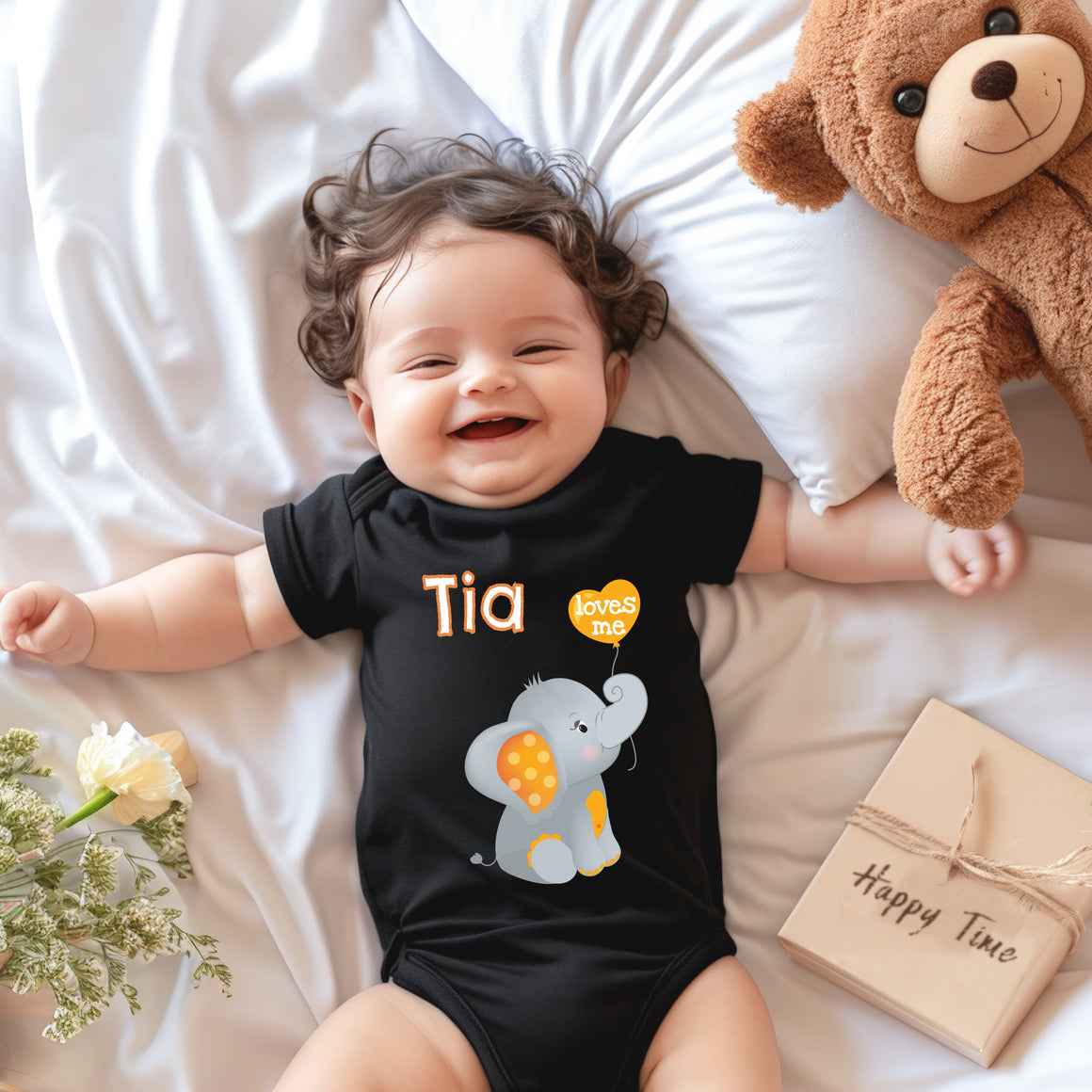 A smiling baby wearing a black t-shirt with an elephant design lies on a bed next to a stuffed teddy bear.