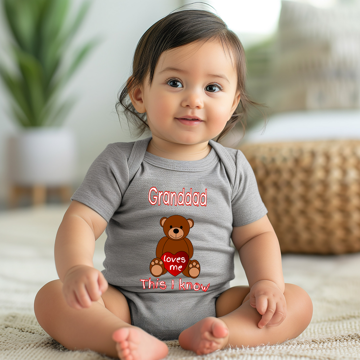 A smiling baby girl wearing a grey onesie with a teddy bear and text that says "Grandpa loves me This I know".