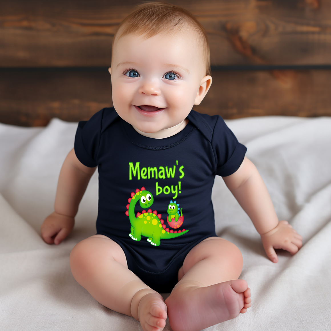 A smiling baby wearing a black shirt with a dinosaur graphic and text that says "Memaw's boy!"
