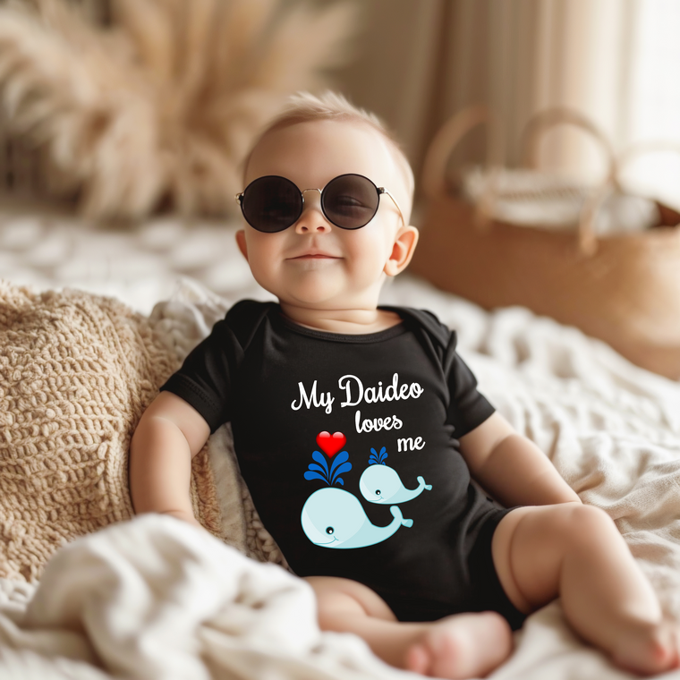 A baby wearing a black onesie with a graphic of a whale and the text "My Daideo loves me" is sitting on a bed.