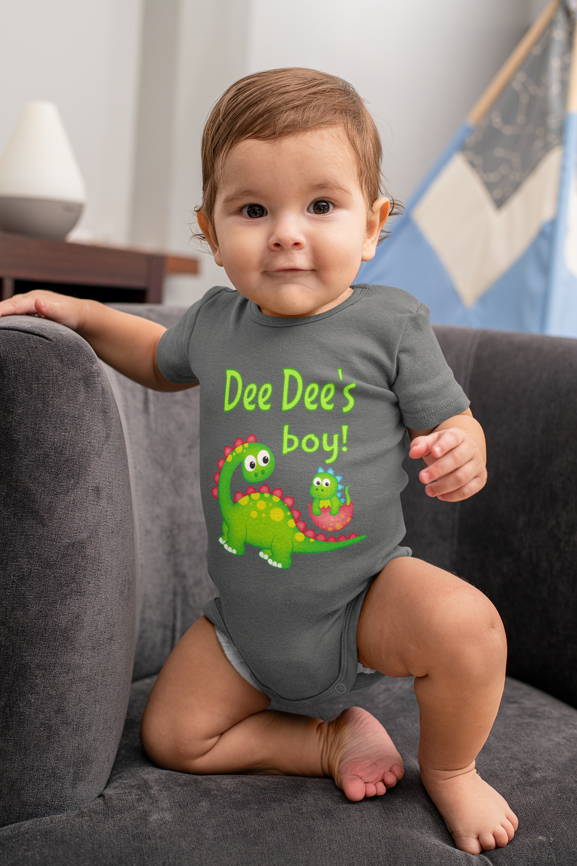 A smiling baby boy wearing a grey onesie with a cartoon dinosaur and the text "Dee Dee's boy!" is sitting on a couch.
