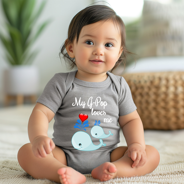 A smiling baby girl wearing a gray onesie with a graphic of a whale and the text "My G-pop loves me" is sitting on the floor.