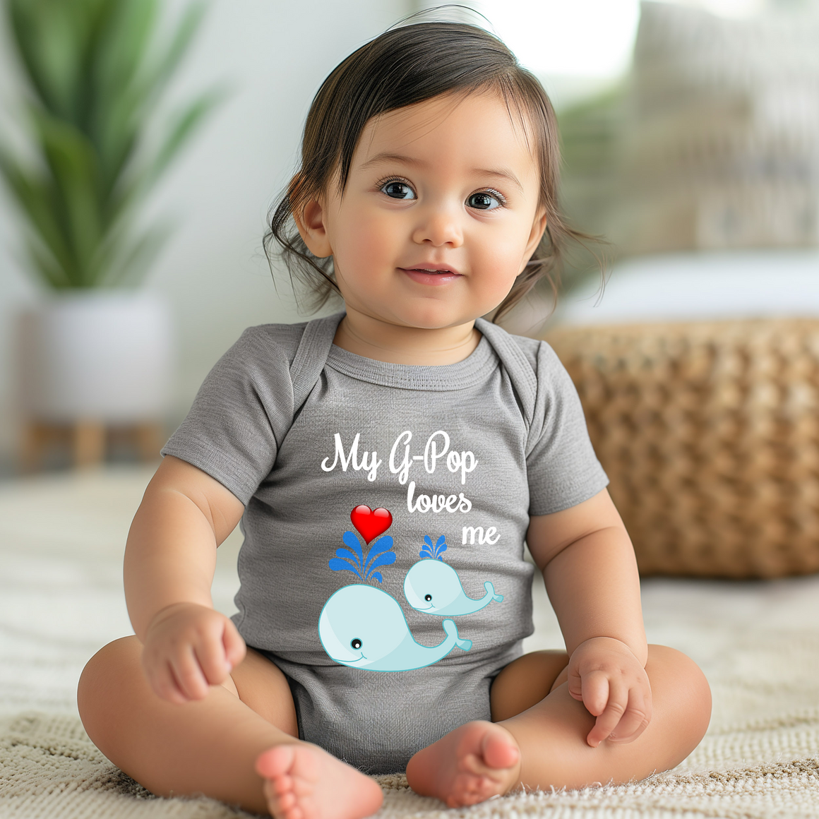 A smiling baby girl wearing a gray onesie with a graphic of a whale and the text "My G-pop loves me" is sitting on the floor.