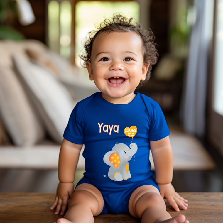A smiling baby boy wearing a blue onesie with an elephant design sits on a wooden surface.