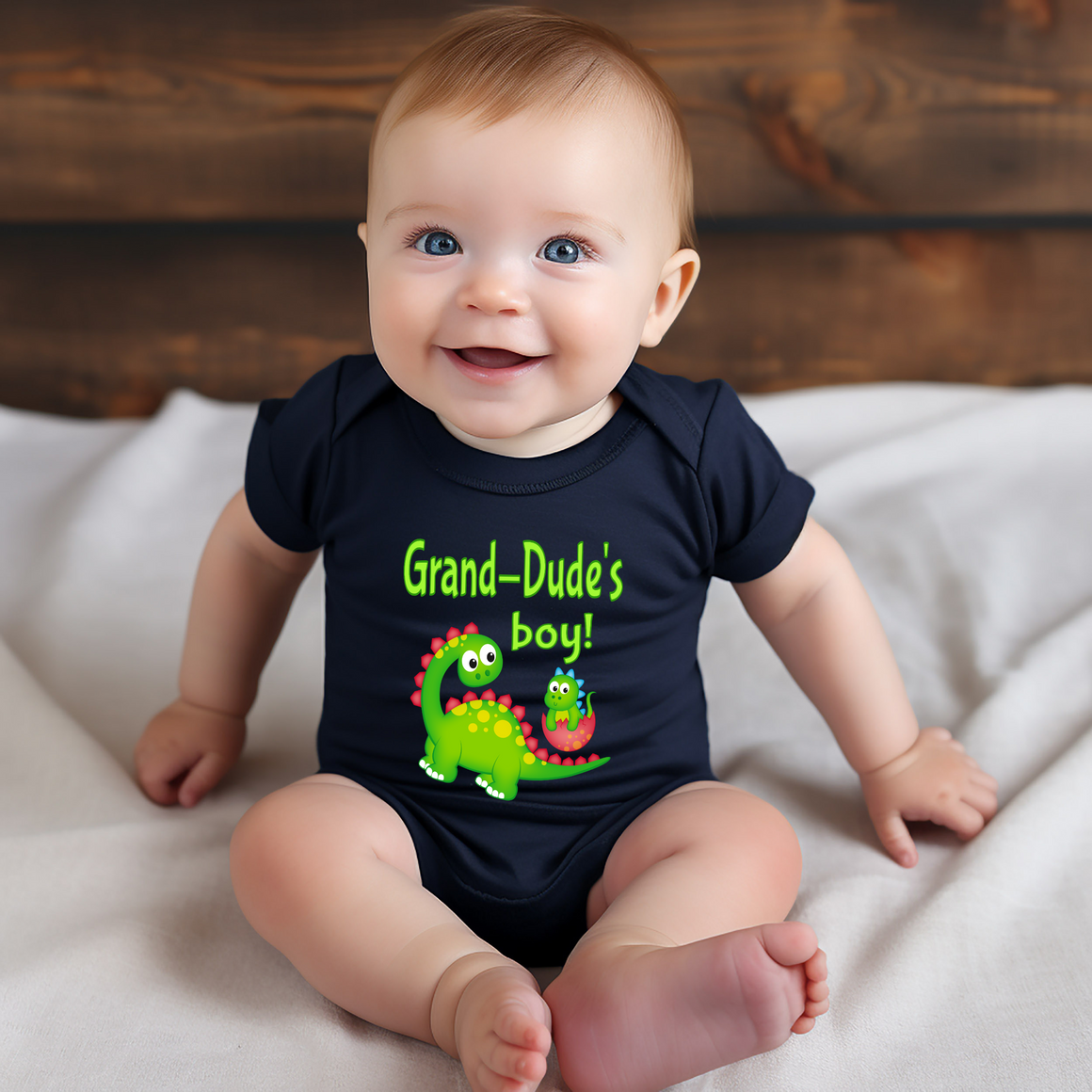 A smiling baby wearing a black shirt with a dinosaur graphic and text that says "Grand-Dude's boy!"