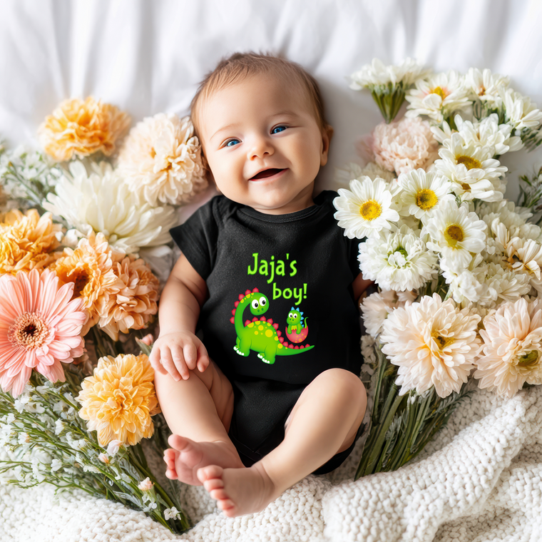 A smiling baby wearing a black onesie with a dinosaur graphic lies on a bed surrounded by colorful flowers.