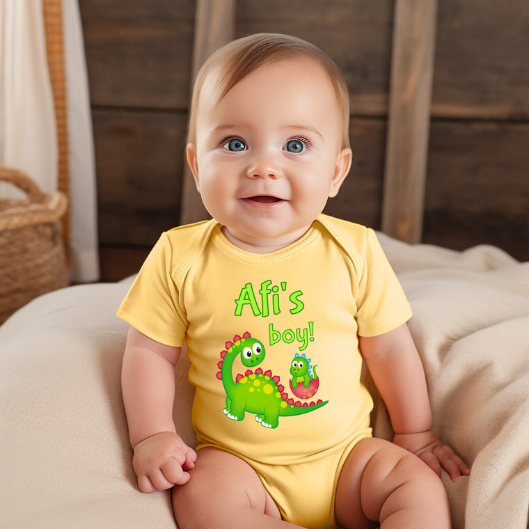 A smiling baby boy wearing a yellow onesie with a dinosaur graphic and the text "Afi's boy!"