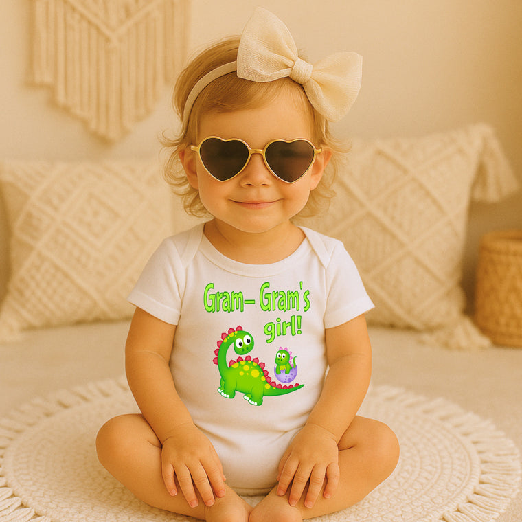 A young girl wearing a white onesie with a dinosaur and the text "Gram- Gram's girl!" sits on a white rug, smiling and wearing heart-shaped sunglasses.