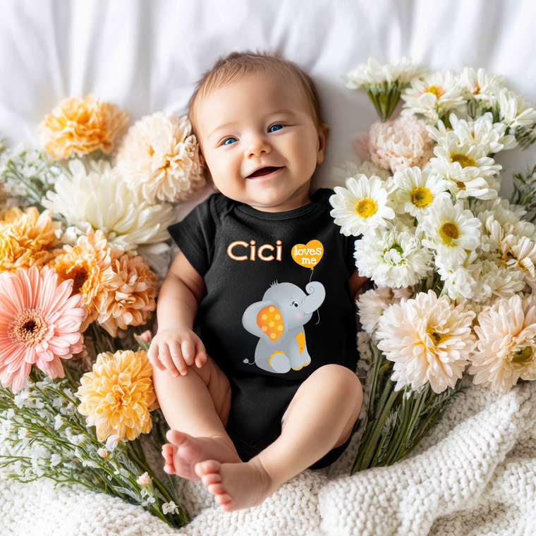 A smiling baby wearing a black outfit with an elephant design lies surrounded by a bouquet of colorful flowers.