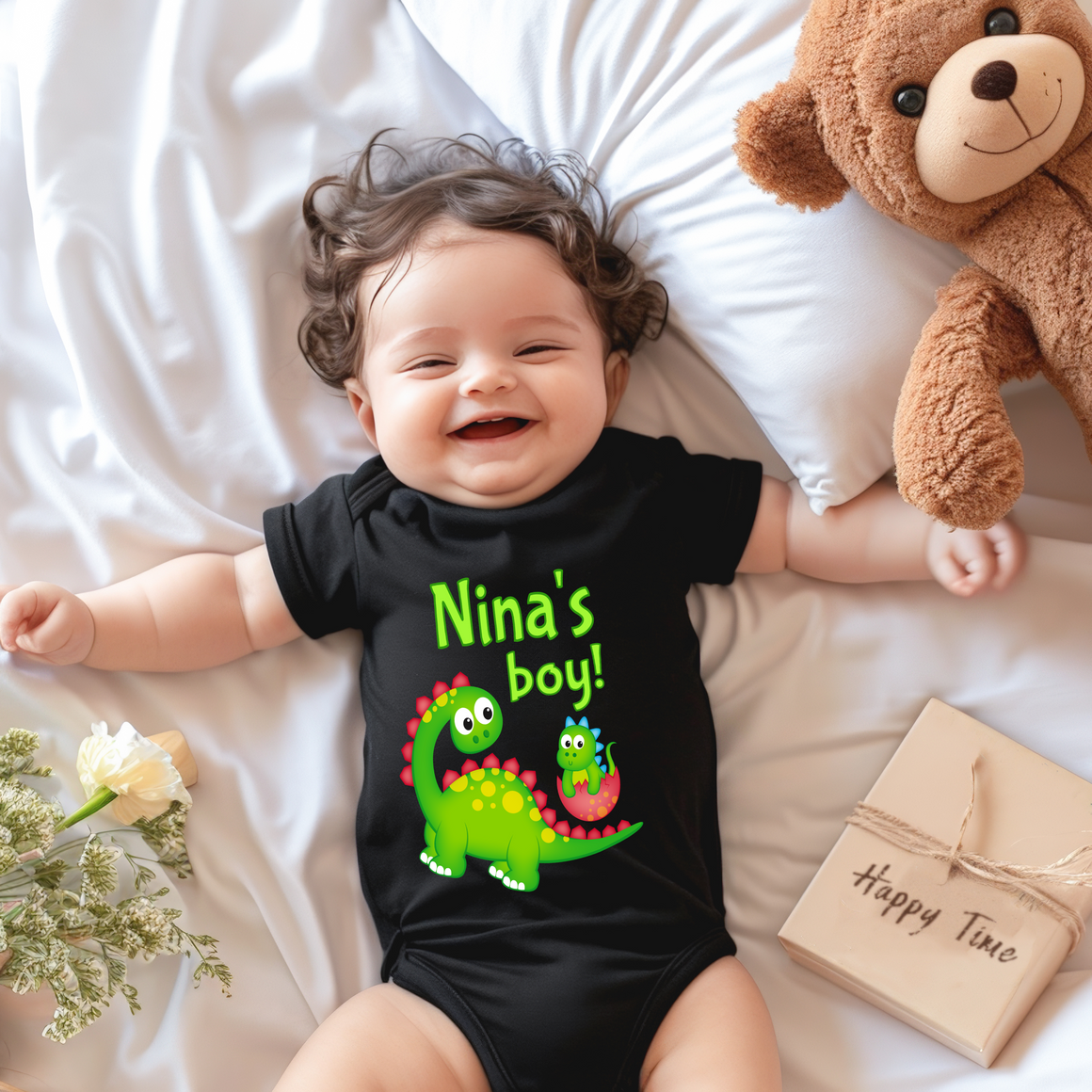 A smiling baby wearing a black onesie with a dinosaur design lies on a bed next to a stuffed teddy bear.