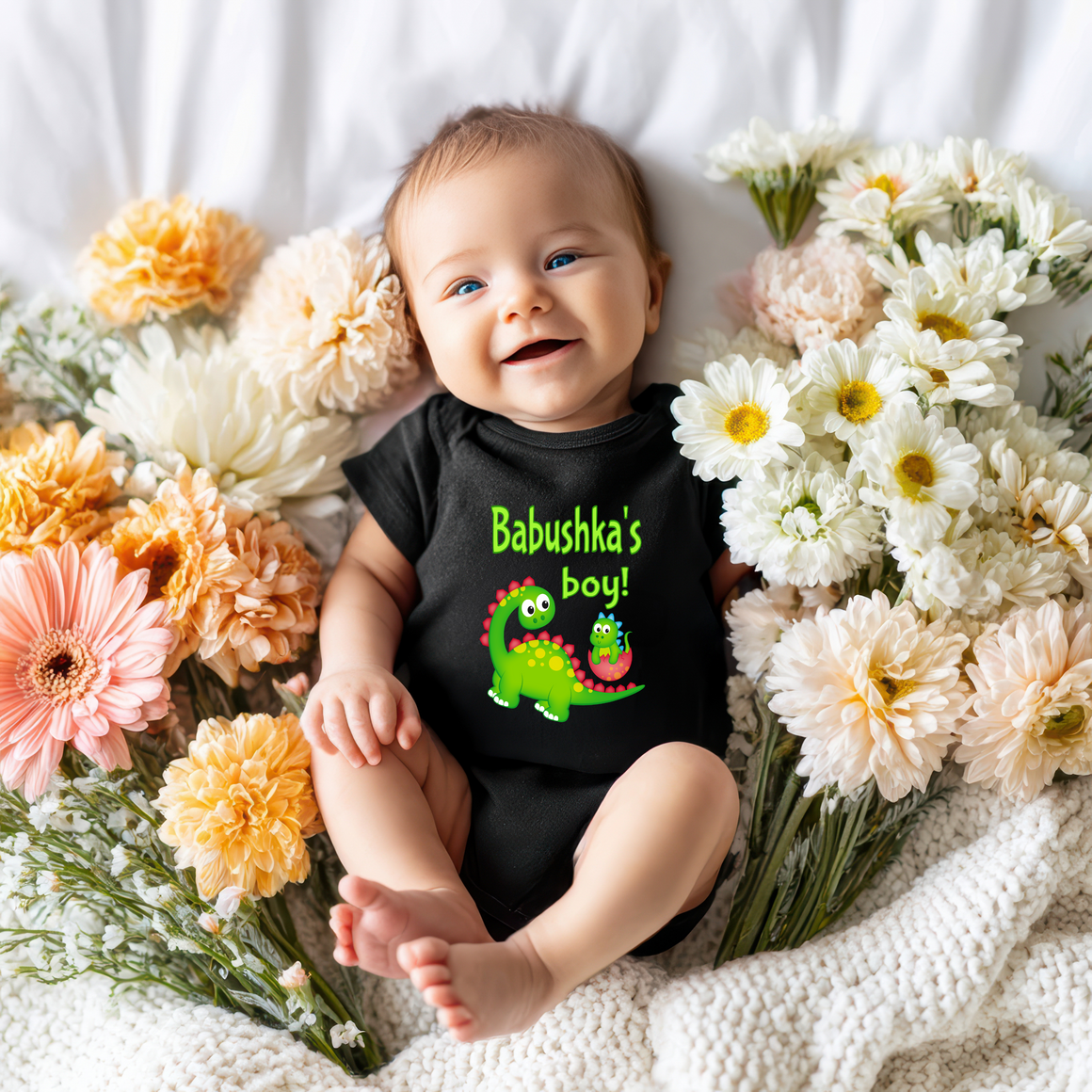 A smiling baby wearing a black onesie with a dinosaur graphic lies on a bed surrounded by colorful flowers.