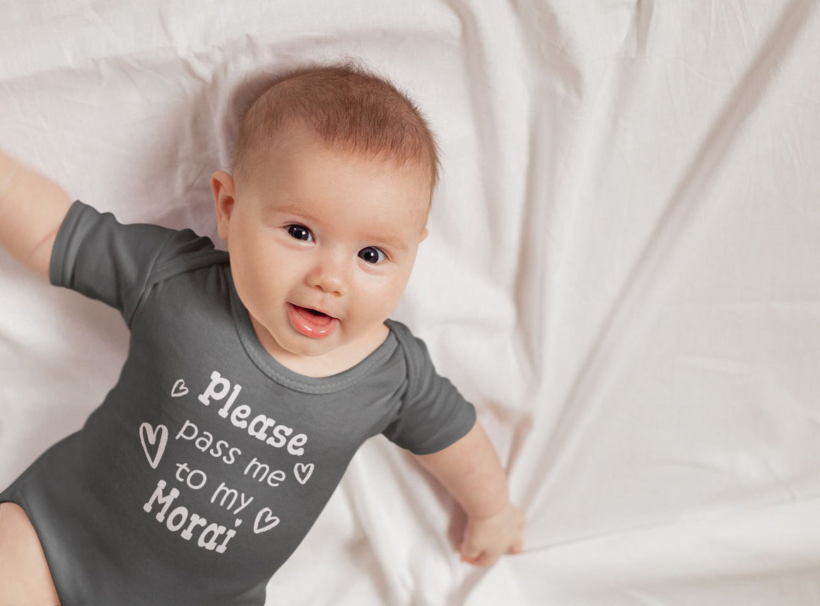 A smiling baby wearing a grey onesie with the text "Please pass me to my mother" lying on a white sheet.