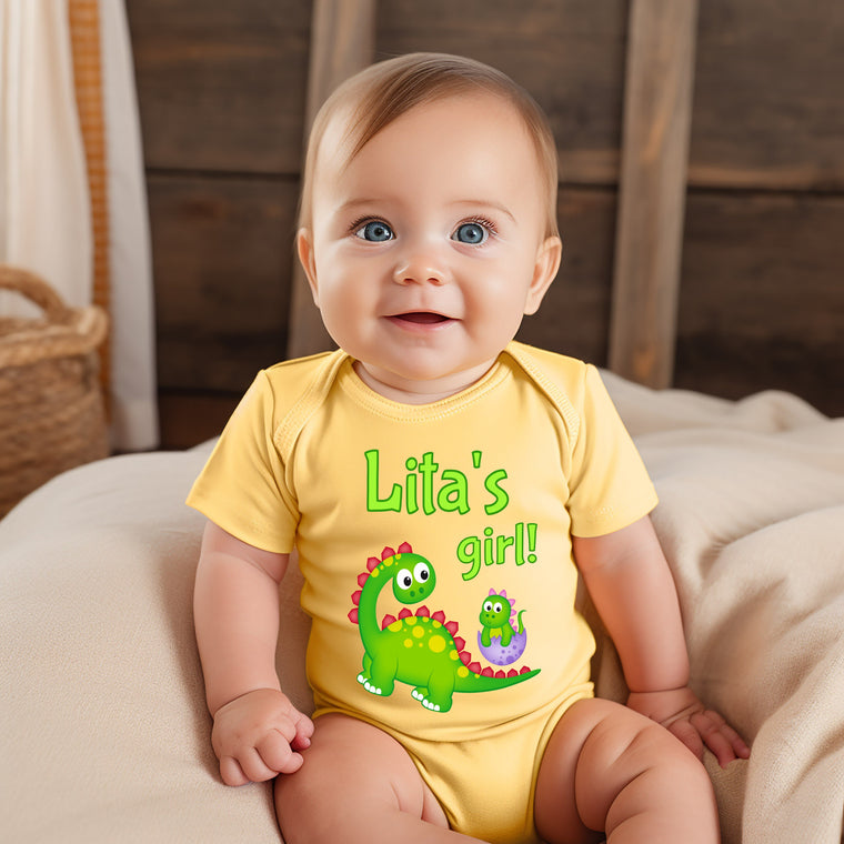 A smiling baby wearing a yellow onesie with a dinosaur design and the text "Lita's girl!"