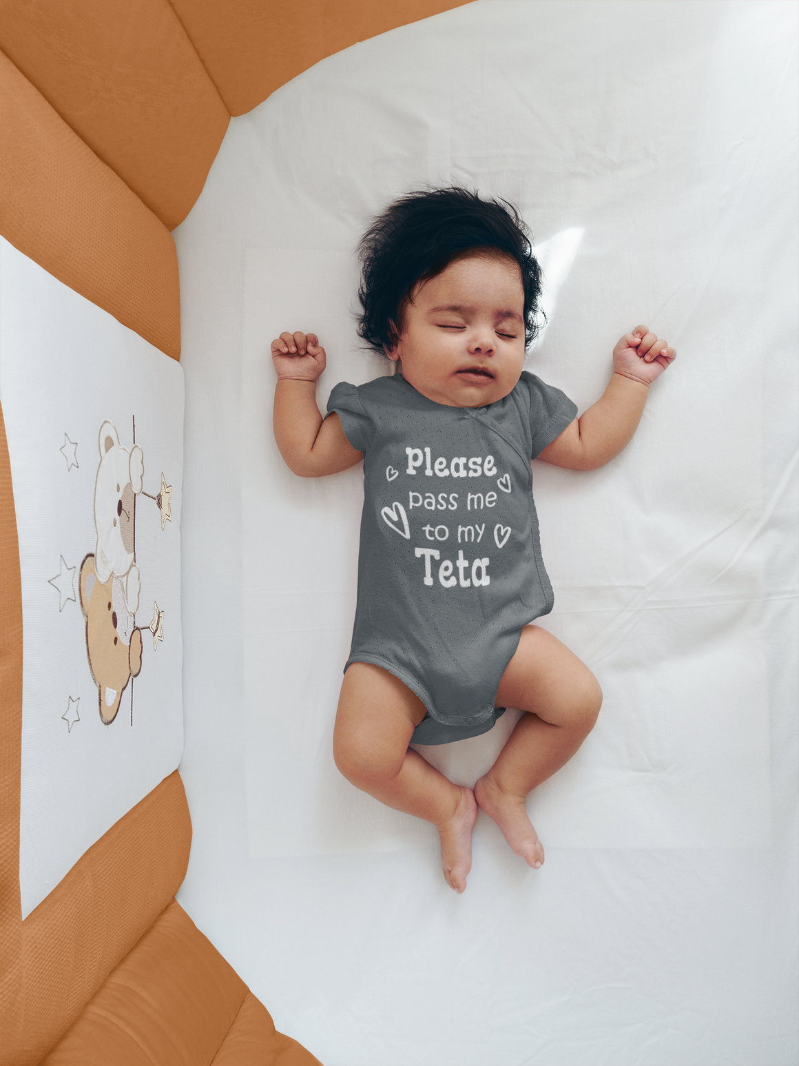 A sleeping baby wearing a gray onesie with the text "Please pass me to my Teta" is lying on a white sheet.