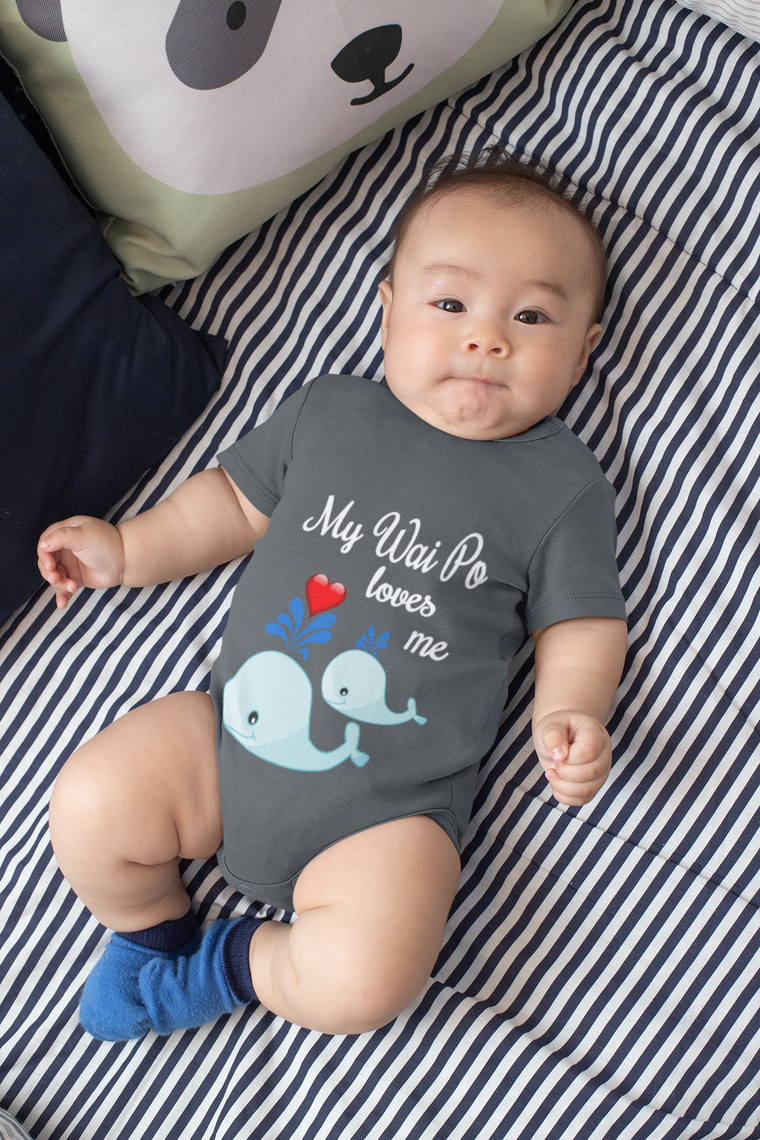 A baby wearing a gray onesie with a design of two blue whale illustrations and the text "My Way To Loves Me" lying on a striped bed.
