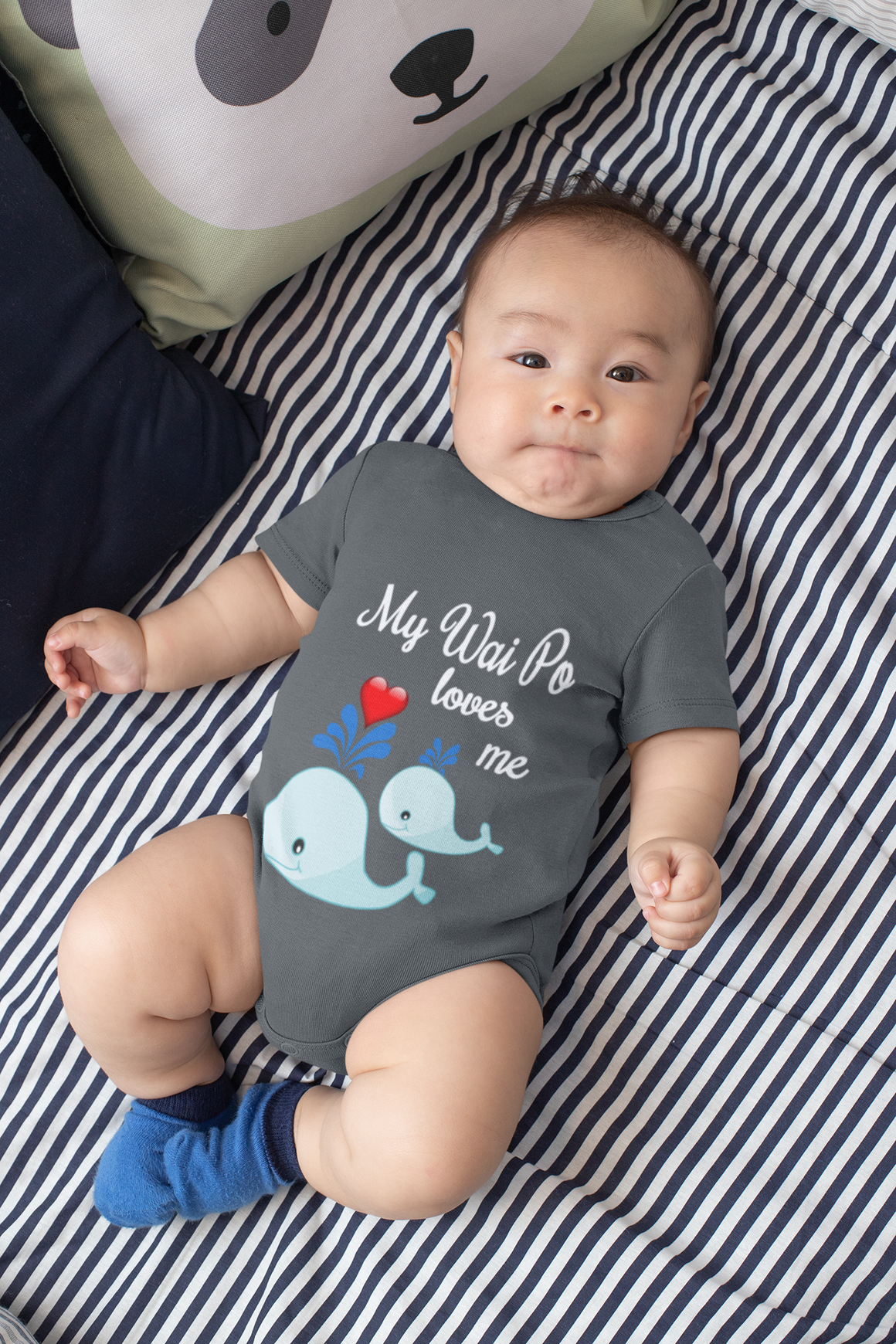 A baby wearing a gray onesie with a design of two blue whale illustrations and the text "My Way To Loves Me" lying on a striped bed.