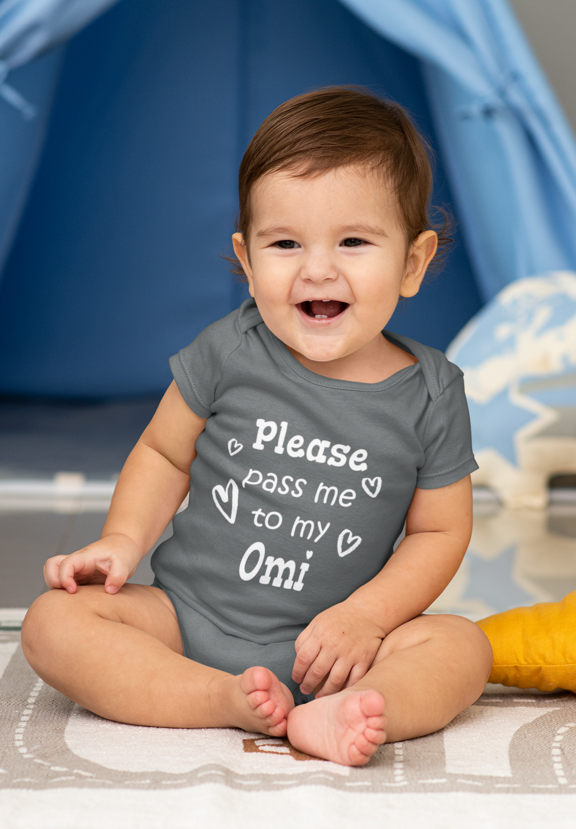 A smiling baby wearing a grey onesie with the text "Please pass me to my Omi" is sitting on a gray and white patterned blanket.
