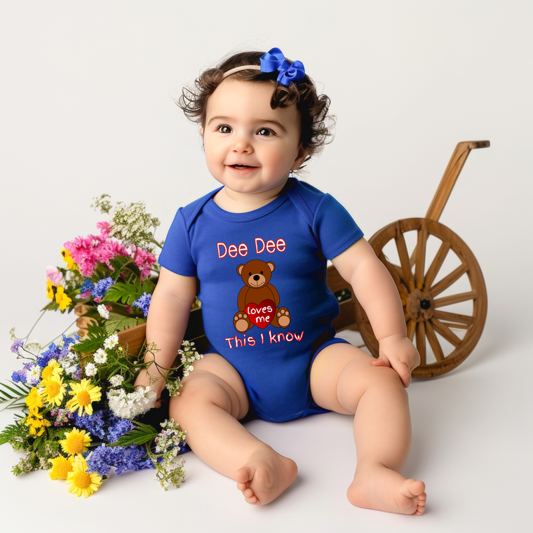 A smiling baby girl wearing a blue onesie with a teddy bear and the text "Dee Dee loves me This I Know" is sitting next to a wooden wagon filled with colorful flowers.