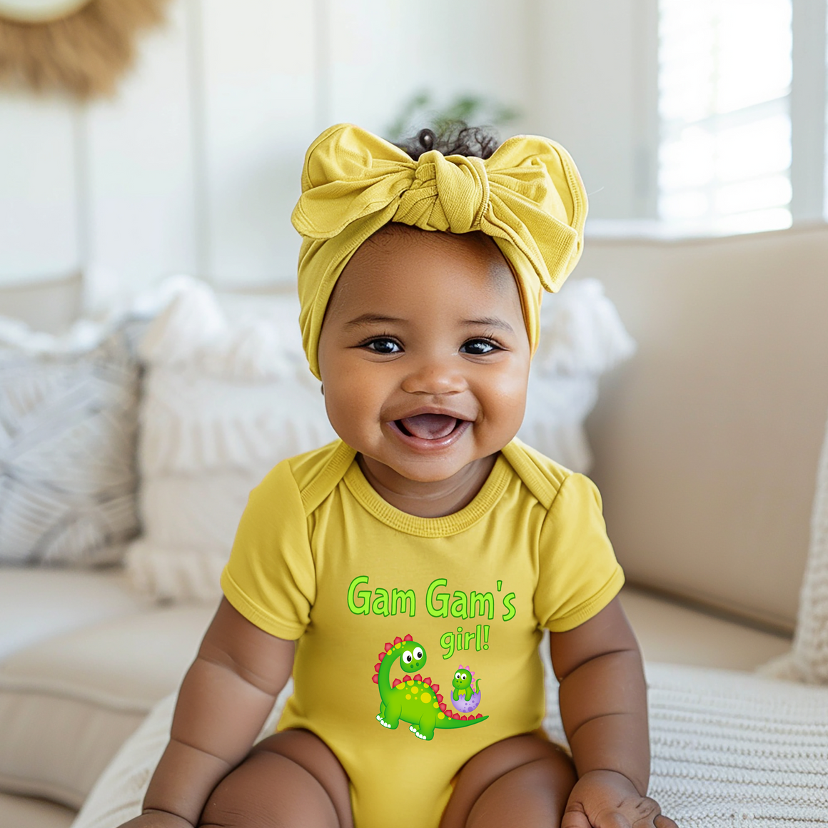 A smiling baby girl wearing a yellow outfit with a dinosaur graphic and a yellow headband sits on a couch.