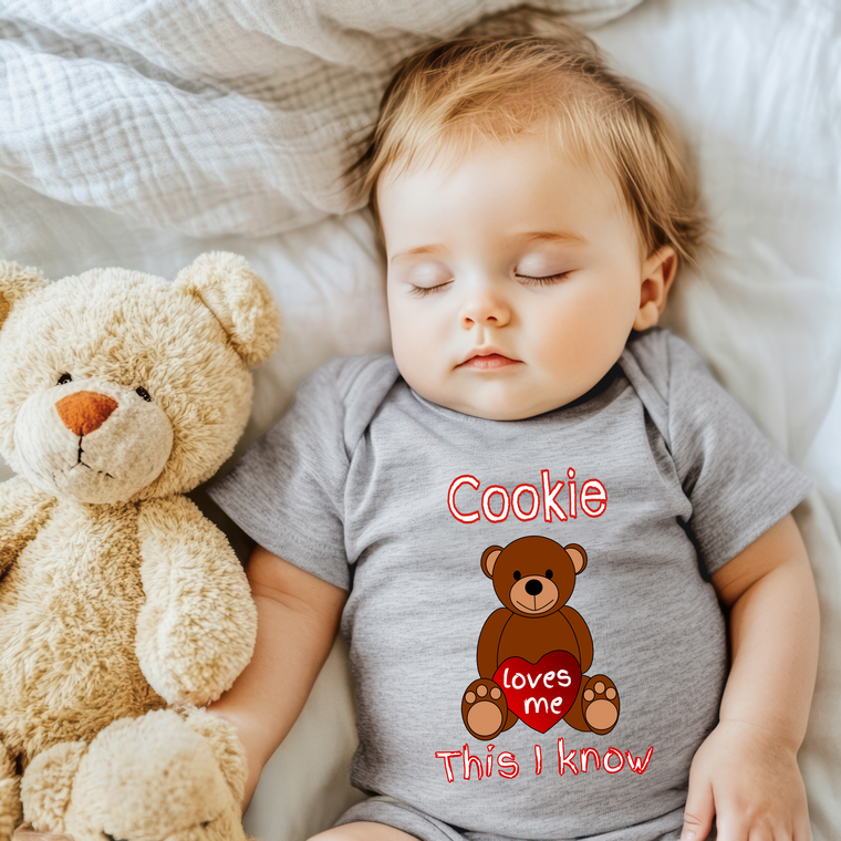 A sleeping baby wearing a gray onesie with a teddy bear and the text "Cookie loves me" printed on it.