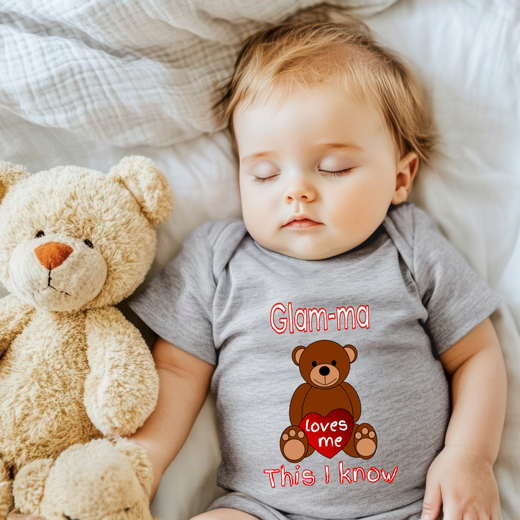 A sleeping baby wearing a gray shirt with a teddy bear and the text "Glam-ma loves me This I know" printed on it.
