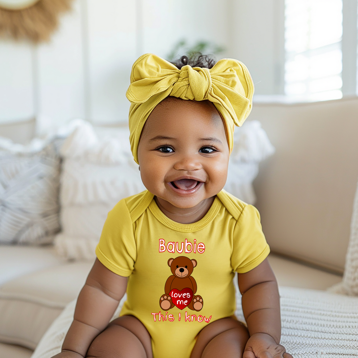 A smiling baby girl wearing a yellow outfit with a teddy bear graphic and the text "Babie loves this I know" is sitting on a couch.