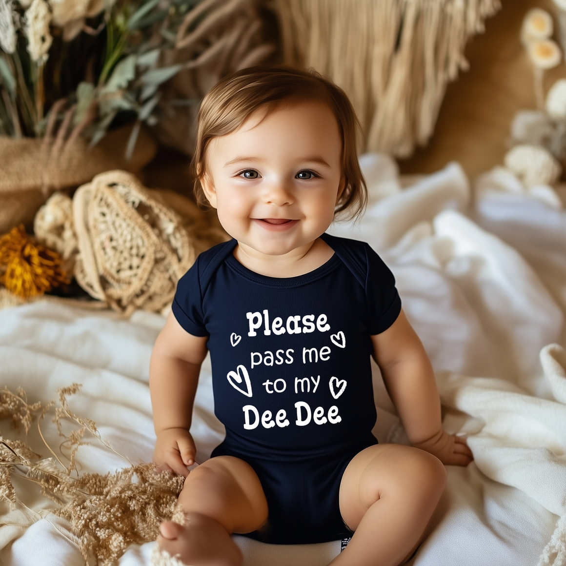 A smiling baby wearing a navy blue onesie with the text "Please pass me to my Dee Dee" sits on a bed with a floral background.