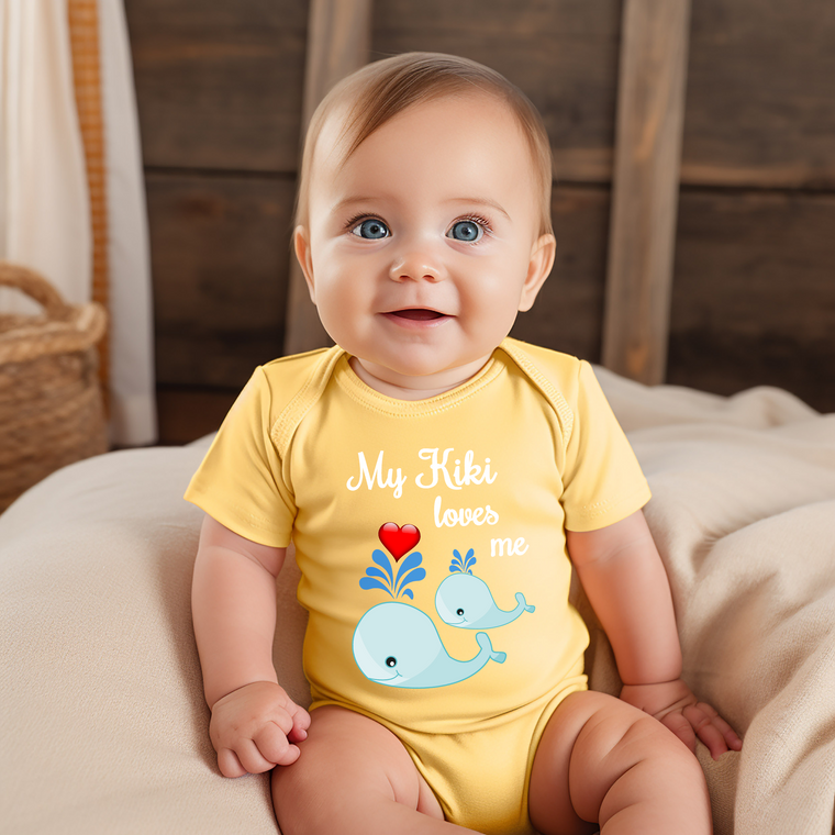 A smiling baby wearing a yellow onesie with a cartoon whale and the text "My Kiki loves me" is sitting on a bed.