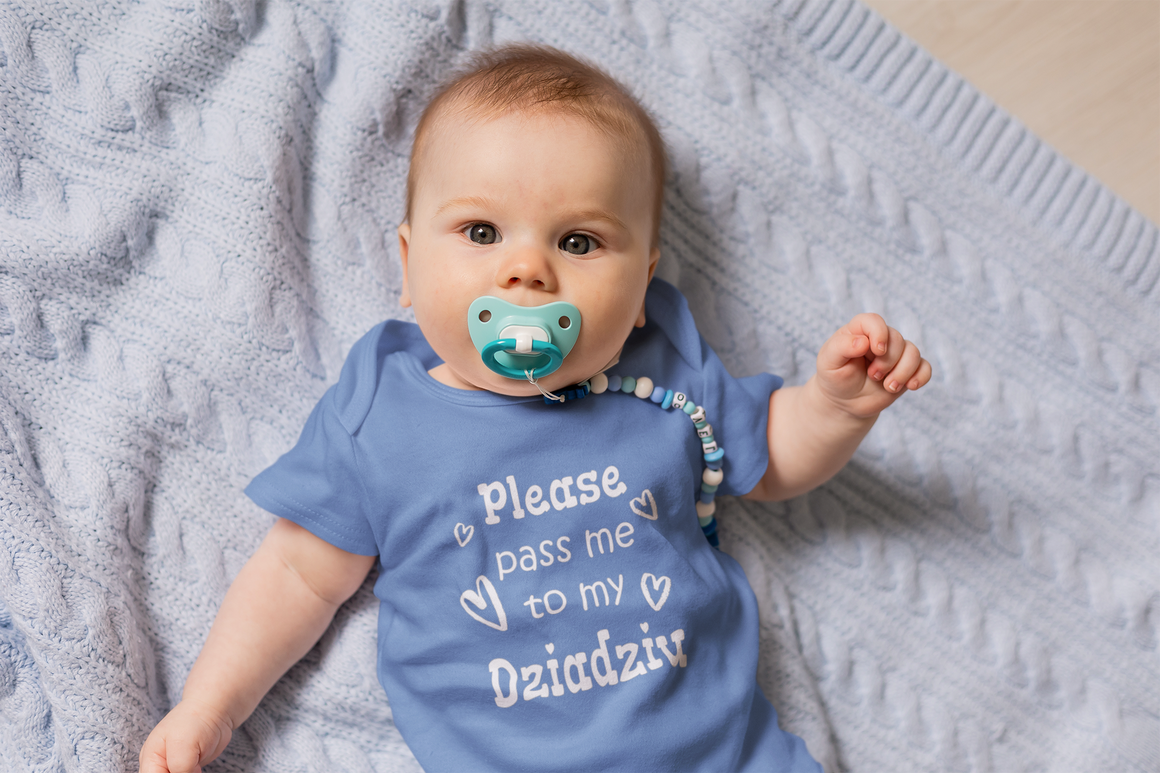 A baby wearing a blue onesie with the text "Please pass me to my Ozadziz" lying on a blue blanket.