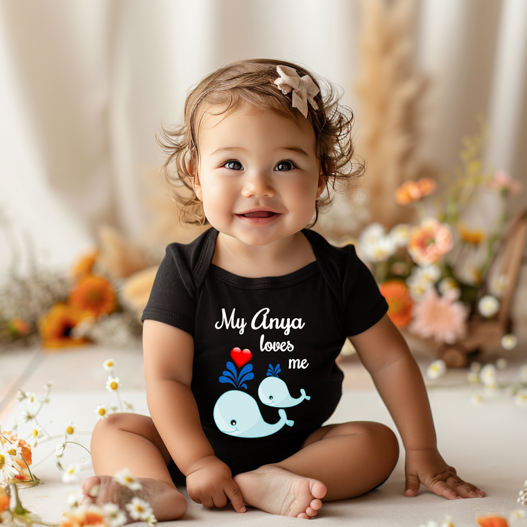 A smiling baby girl wearing a black shirt with a whale graphic and the text "My Anya loves me" is sitting on the floor surrounded by flowers.