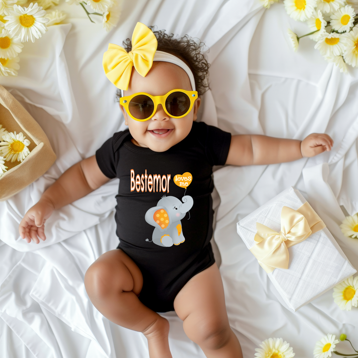 A smiling baby girl wearing a black outfit with a yellow bow and sunglasses lies on a white sheet, surrounded by yellow flowers.