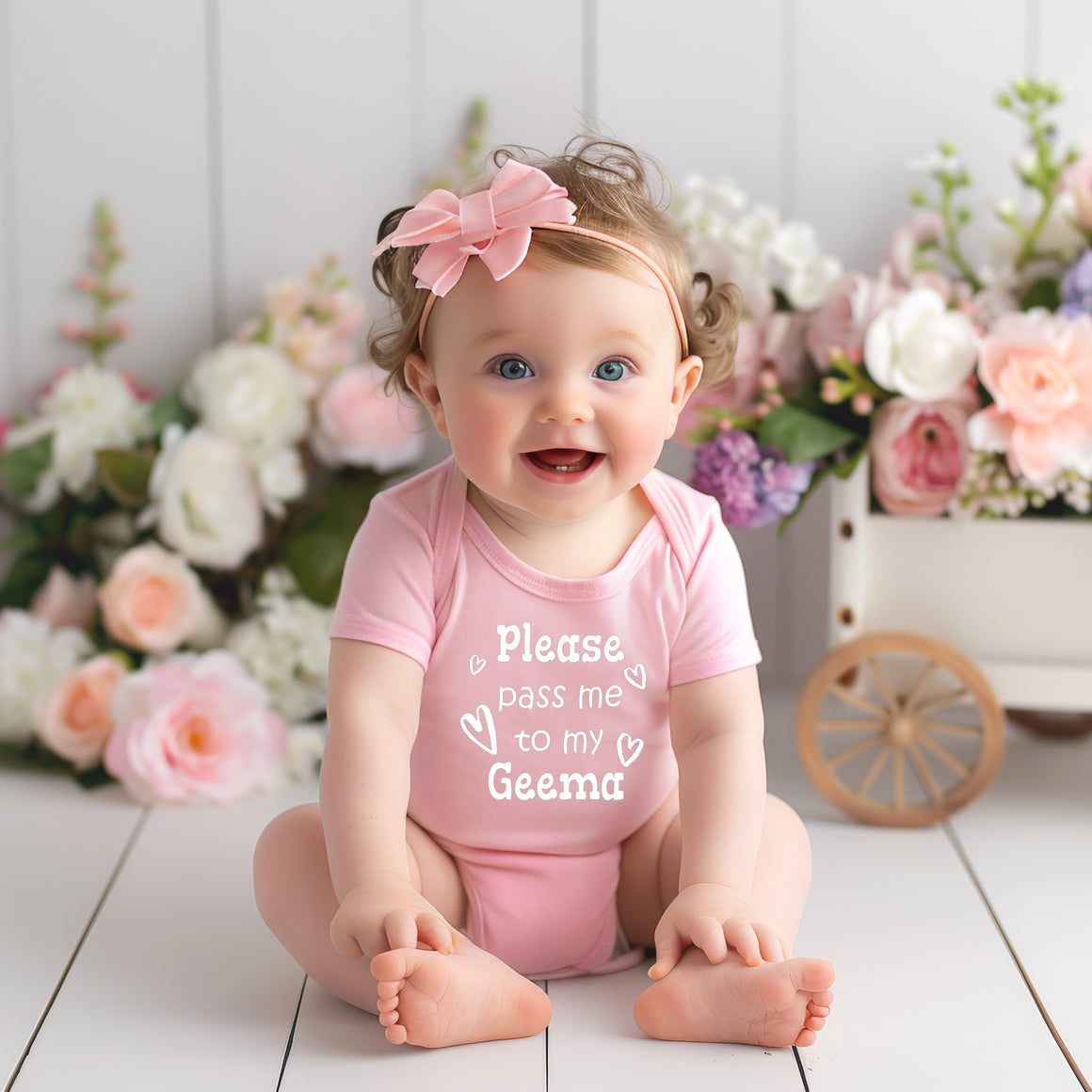 A smiling baby girl wearing a pink outfit with a bow sits in front of a floral background.