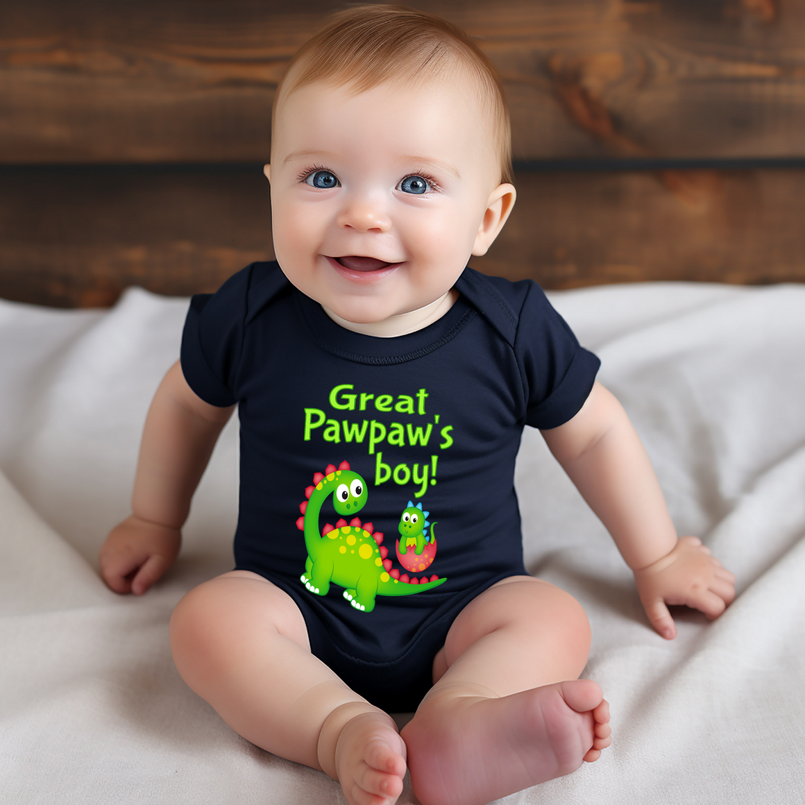 A smiling baby wearing a black shirt with a dinosaur graphic and text that says "Great Pawpaw's boy!"