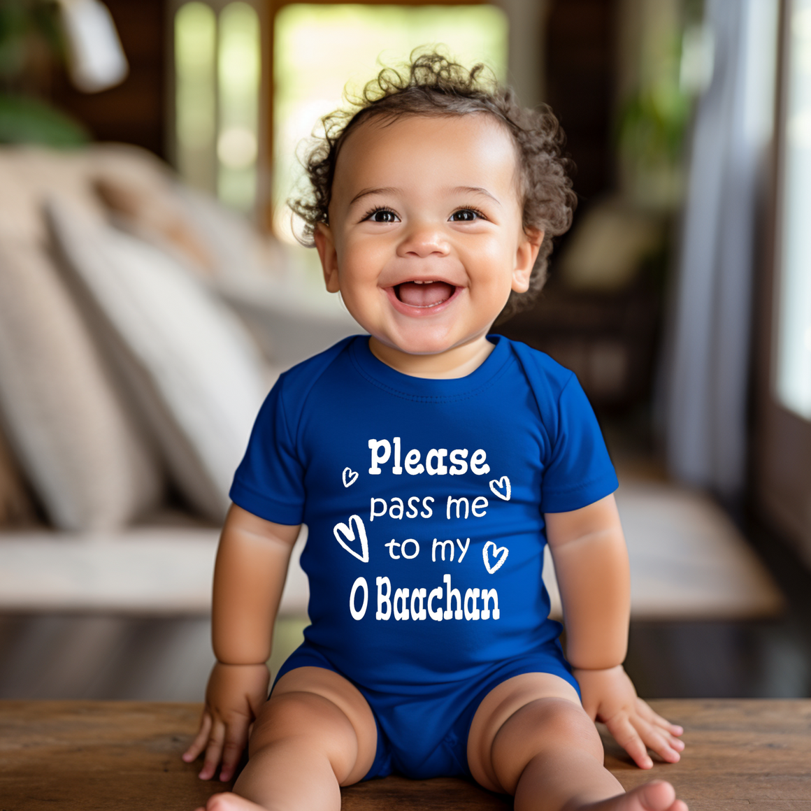 A smiling baby boy wearing a blue onesie with the text "Please pass me to my O Baachan" is sitting on a wooden surface.
