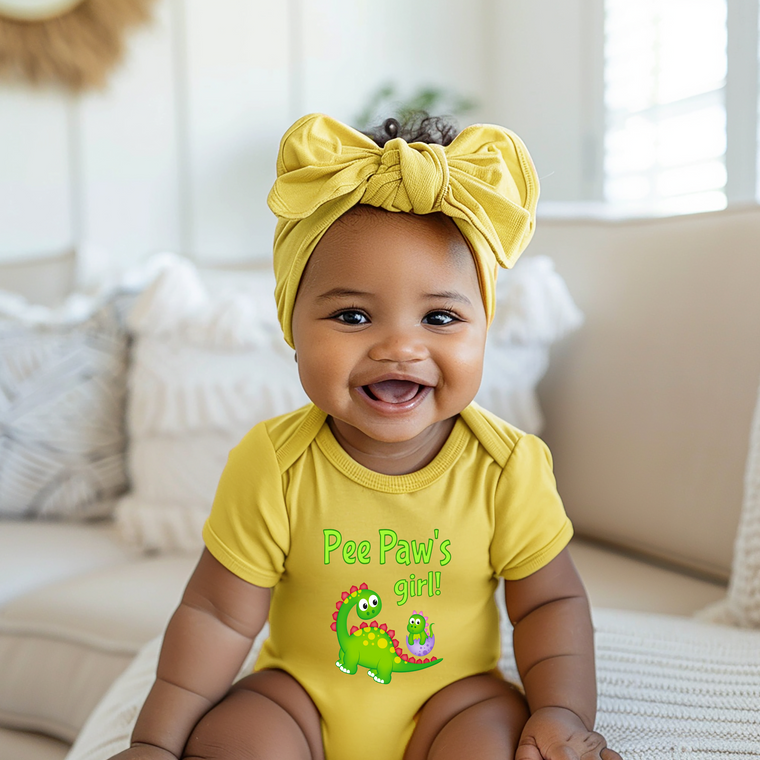 A smiling baby girl wearing a yellow outfit with a bow and a green dinosaur on it, sitting on a couch.
