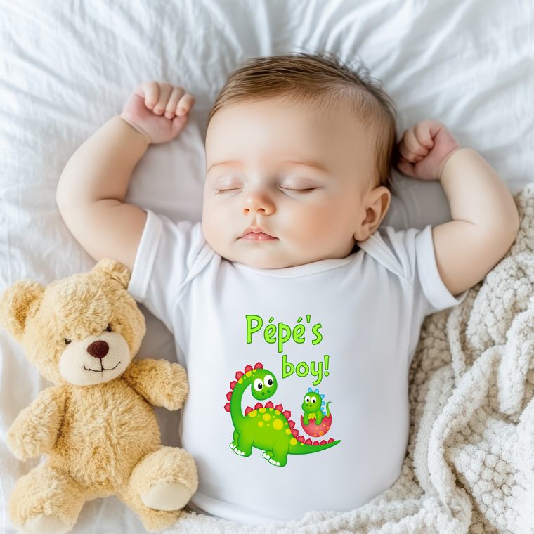 A sleeping baby wearing a white onesie with a dinosaur graphic and the text "Péppe's boy!" next to a teddy bear.