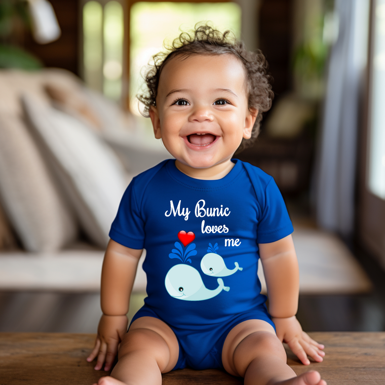 A smiling baby boy wearing a blue onesie with a whale design and the text "My Bunic loves me" is sitting on a wooden surface.