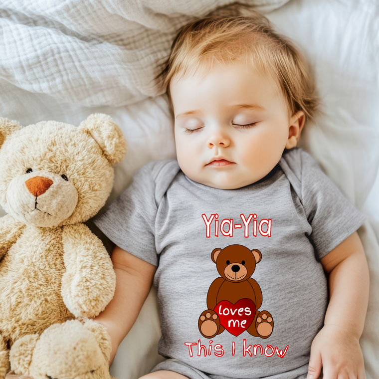 A sleeping baby wearing a gray onesie with a teddy bear and the text "Yia-Yia loves me This I know" printed on it.