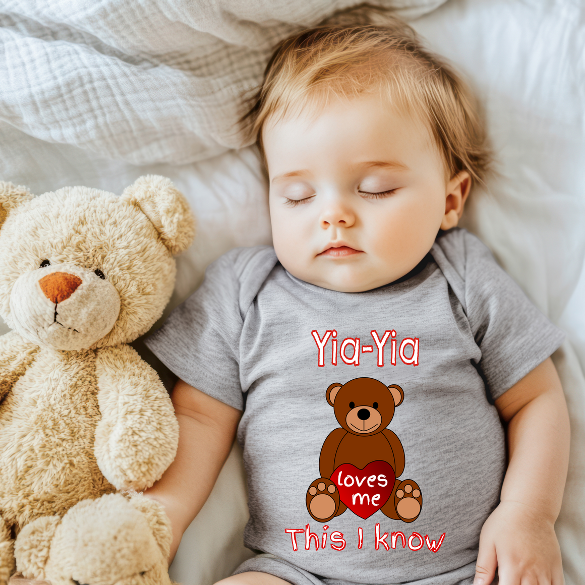 A sleeping baby wearing a gray onesie with a teddy bear and the text "Yia-Yia loves me This I know" printed on it.