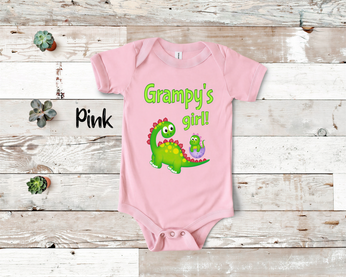 A pink baby onesie with a green dinosaur and the text "Grampy's girl!" printed on it, placed on a wooden surface.