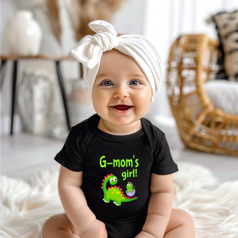 A smiling baby girl wearing a black t-shirt with a dinosaur graphic and the text "G-mom's girl!"