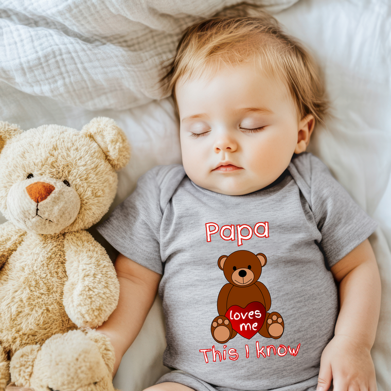 A sleeping baby wearing a gray onesie with a teddy bear and the text "Papa loves me" printed on it.