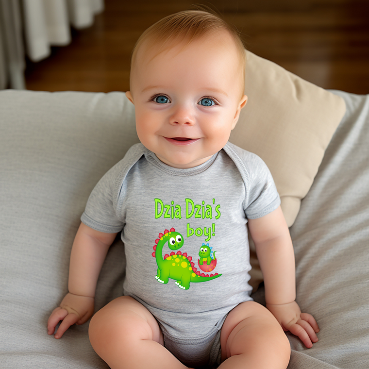A smiling baby boy wearing a gray onesie with a dinosaur graphic and the text "Dzia Dzia's boy!"
