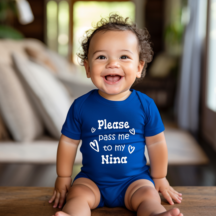 A smiling baby boy wearing a blue onesie with the text "Please pass me to my NINA" is sitting on a wooden surface.