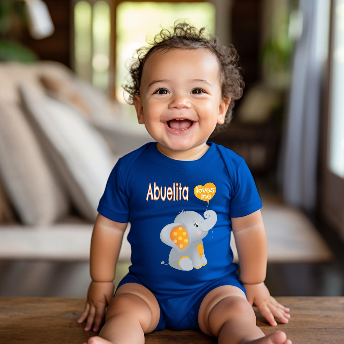 A smiling baby boy wearing a blue onesie with an elephant design sits on a wooden surface.