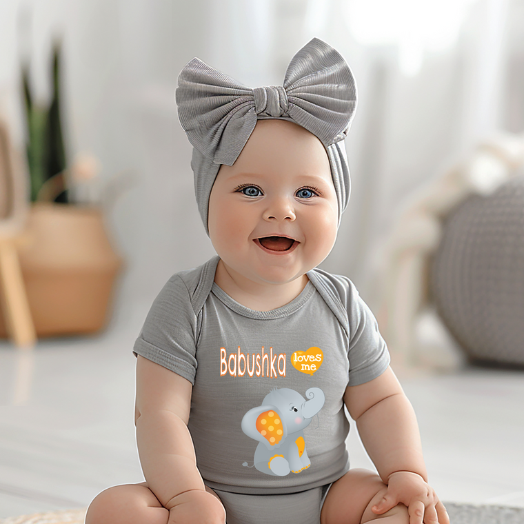 A smiling baby girl wearing a gray headband and a gray onesie with an elephant design, sitting on a white floor.